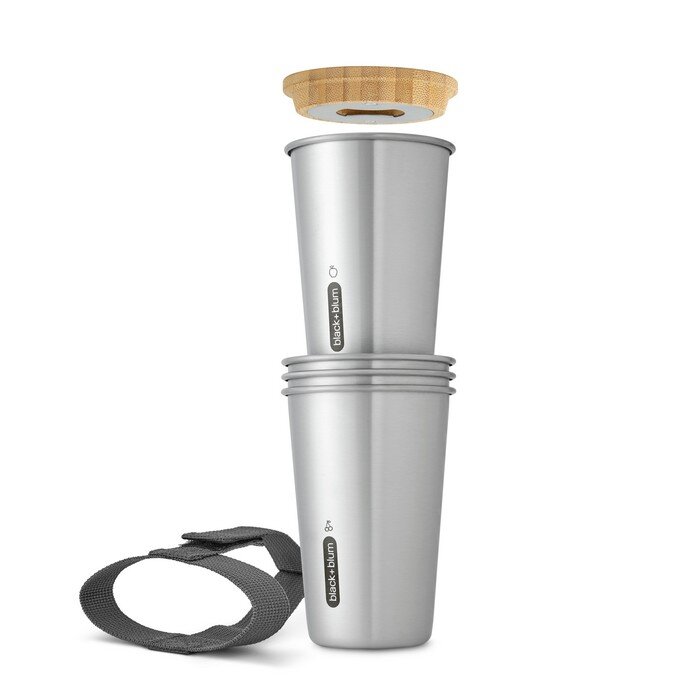 Na obrazie widoczne są dwa stalowe kubki ustawione jeden na drugim, z logo \'Black+Blum\'. Nad nimi znajduje się drewniana pokrywka, a obok leży szara opaska.