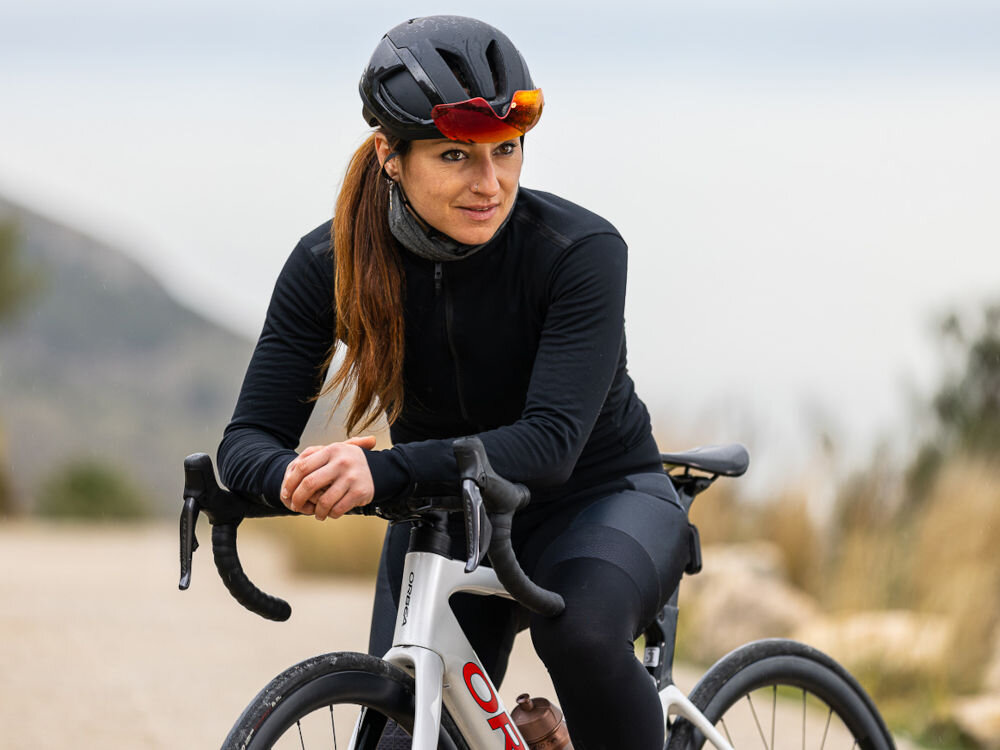 Kask rowerowy LAZER Vento KinetiCore Kobieta w czarnym stroju kolarskim i kasku szosowym Lazer Vento KinetiCore oparta o kierownicę roweru Orbea.