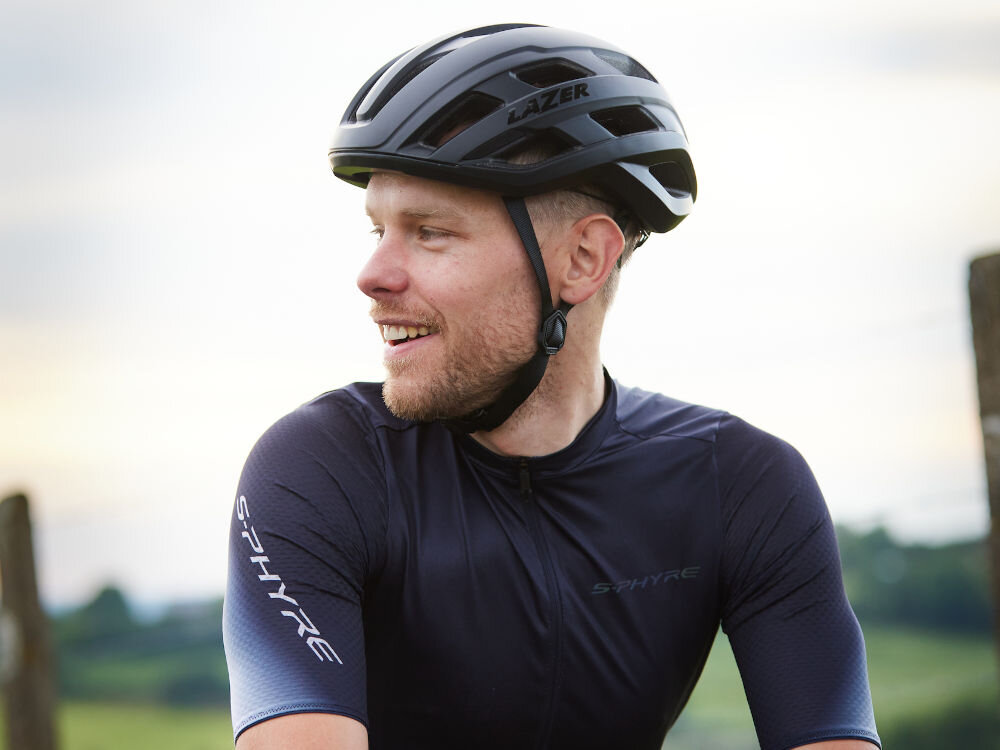 Kask rowerowy LAZER Strada KinetiCore Czarny Szosowy (rozmiar S) osoba w kasku siedzi na rowerze patrzy w dal widok bokiem w tle łąka pole kolarze szosowi lekka konstrukcja z poliwęglanu wewnętrzna warstwa EPS skuteczna ochrona przy zachowaniu wygody użytkowania strefa kontrolowanego zgniotu ogranicza ryzyko kontuzji poczucie kontroli podczas jazdy