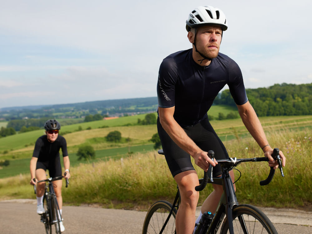 Kask rowerowy LAZER Strada KinetiCore Biały Szosowy (rozmiar L) droga ulica szosa dwie osoby w kaskach jadna na rowerach po drodze w tle pola łąki las bezpieczeństwo komfort wsparcie