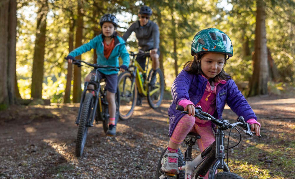 Kask rowerowy LAZER Nutz KinetiCore 2.0 Zielony dla Dzieci (rozmiar 50-56) park las drzewa na pierwszym planie dziecko w kasku jedzie przed siebie w tle dwie osoby w kaskach jadą na rowerach bezpieczeństwo i komfort najmłodszych użytkowników chroni głowę podczas upadków i uderzeń lekki i wygodny w noszeniu regulowane paski siateczka przeciw owadom osłona skroni ochrona i komfort