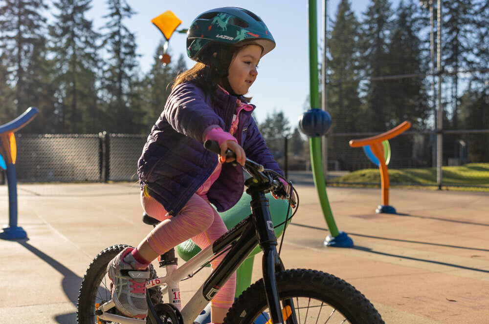 Kask rowerowy LAZER Nutz KinetiCore 2.0 Zielony dla Dzieci (rozmiar 50-56) plac zabaw dziecko w kasku jeździ na rowerze w tle drzewa niebo drabinki dodatkowa osłona skroni zwiększa ochronę w newralgicznych miejscach głowy ryzyko urazów podczas upadku lub uderzenia jest znacząco zmniejszone bezpieczeństwo użytkownika lekka konstrukcja z poliwęglanu