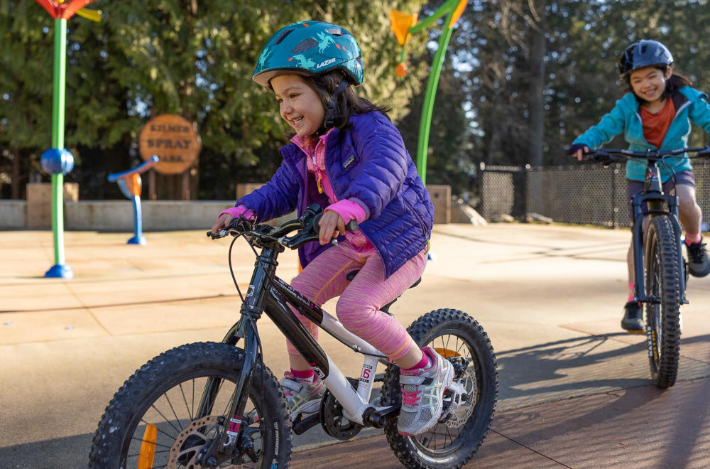 Kask rowerowy LAZER Nutz KinetiCore 2.0 Zielony dla Dzieci (rozmiar 50-56) park plac zabaw dwójka dzieci w kaskach jeżdżą na rowerach w tle drzewa wnętrze kasku wyposażono w miękką wyściółkę materiał EPS skutecznie pochłania energię waga około 240 g homologacje CE EN 1078 i CPSC spełnienie norm bezpieczeństwa kask został przetestowany pod kątem realnych zagrożeń zapewnia ochronę na poziomie wymaganym podczas jazdy po drogach publicznych