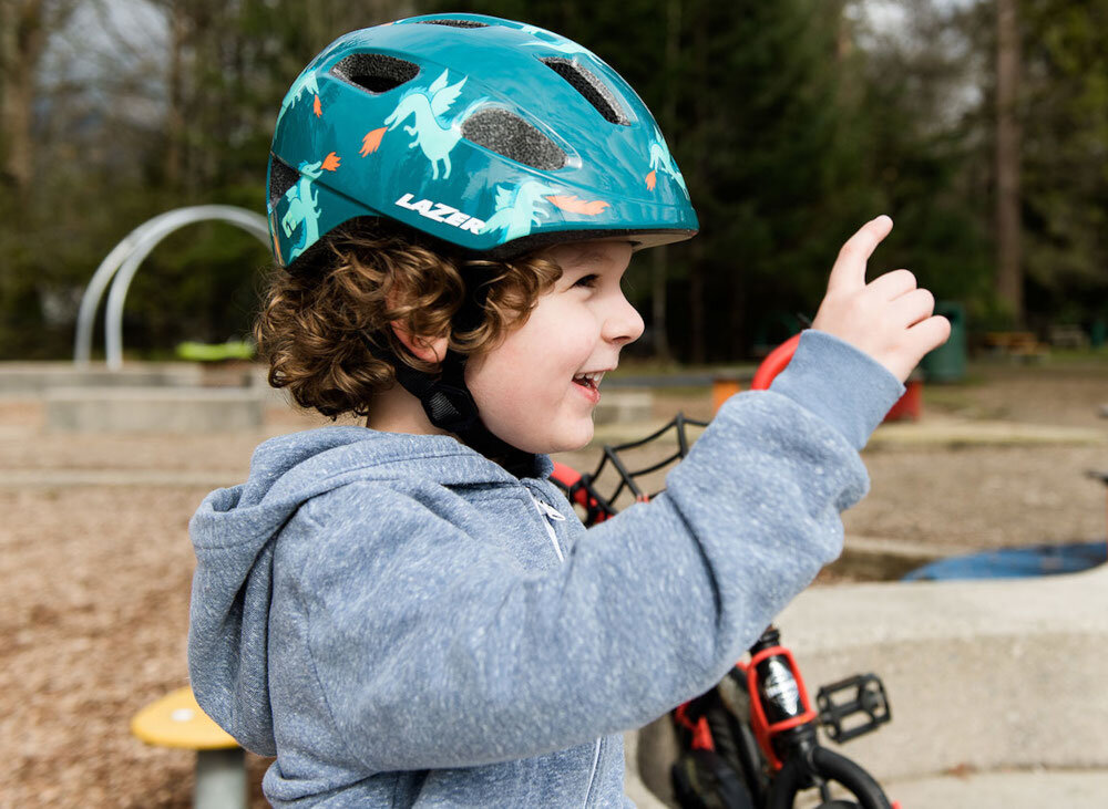 Kask rowerowy LAZER Nutz KinetiCore 2.0 Zielony dla Dzieci (rozmiar 50-56) park plac zabaw dziecko w kasku patrzy przed siebie w tle huśtawki drzewa klamra w kasku została sprytnie umieszczona na policzku eliminuje ryzyko bolesnego szczypania skóry podczas zapinania dzieci mogą łatwo i szybko dopasować kask komfort codziennego użytkowania pozytywne nastawienie do noszenia kasku wspiera bezpieczne poruszanie się