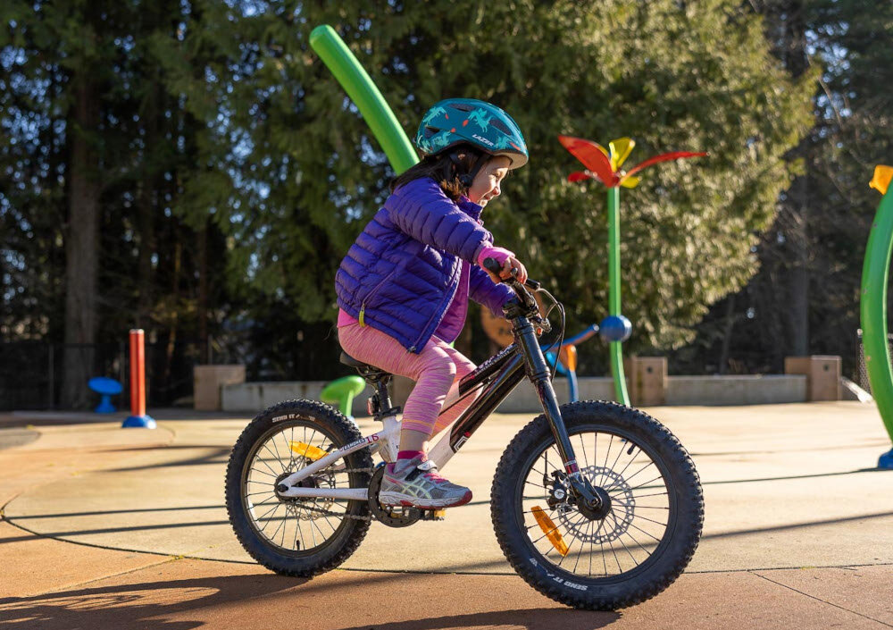 Kask rowerowy LAZER Nutz KinetiCore 2.0 Zielony dla Dzieci (rozmiar 50-56) park plac zabaw dziecko w kasku jedzie na rowerze po drodze w tle drzewa wentylacyjne 12 przemyślanych otworów wentylacyjnych wspierają efektywny obieg powietrza wewnątrz kasku dziecko odczuwa mniejsze nagrzewanie głowy równomierne chłodzenie bez utraty bezpieczeństwa spokojna i przyjemna jazda