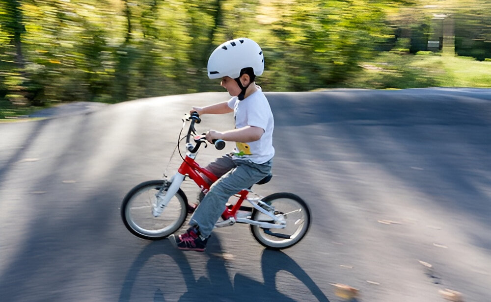 Kask rowerowy LAZER Maze Jr KinetiCore Biały dla Dzieci(rozmiar S/M) droga ulica dziecko w kasku jedzie na rowerze w tle drzewa krzewy bezpieczeństwo komfort wsparcie