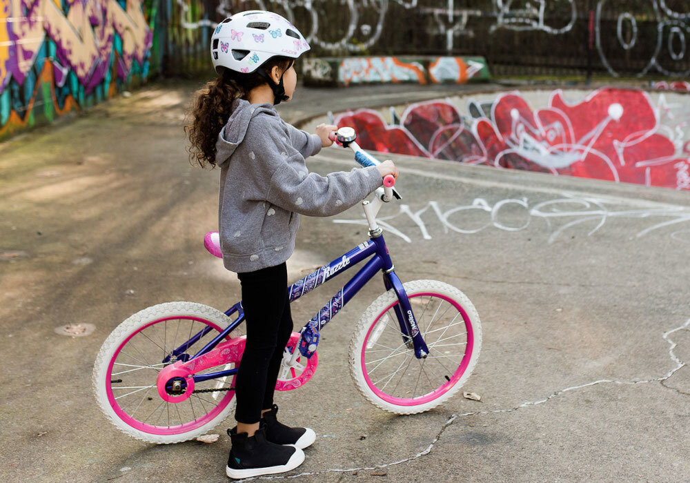 Kask rowerowy LAZER Nutz KinetiCore 2.0 Biały dla Dzieci (rozmiar 50-56) skatepark dziecko w kasku siedzi na rowerze w tle ściana skateparku dziecko widoczne od tyłu dodatkowa osłona skroni zwiększa ochronę w newralgicznych miejscach głowy ryzyko urazów podczas upadku lub uderzenia jest znacząco zmniejszone bezpieczeństwo użytkownika lekka konstrukcja z poliwęglanu