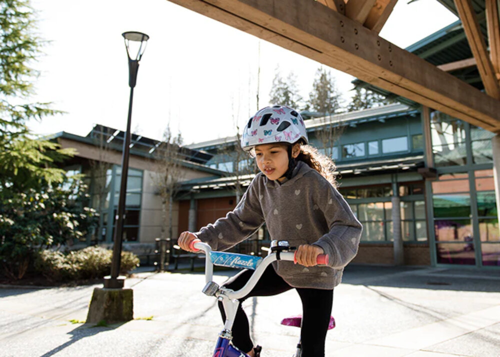 Kask rowerowy LAZER Nutz KinetiCore 2.0 Biały dla Dzieci (rozmiar 50-56) ulica dziecko w kasku siedzi na rowerze w tle budynek lampa niebo wnętrze kasku wyposażono w miękką wyściółkę materiał EPS skutecznie pochłania energię waga około 240 g homologacje CE EN 1078 i CPSC spełnienie norm bezpieczeństwa kask został przetestowany pod kątem realnych zagrożeń zapewnia ochronę na poziomie wymaganym podczas jazdy po drogach publicznych