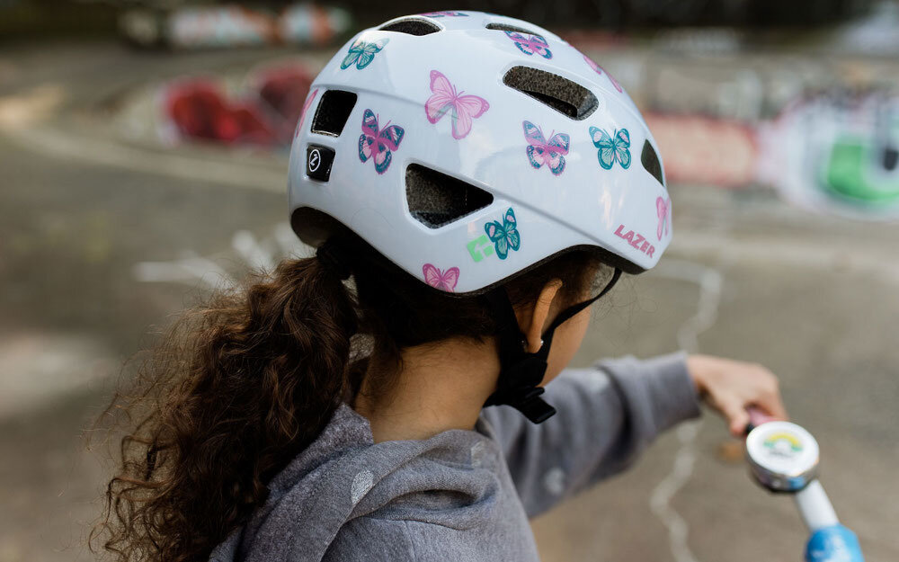 Kask rowerowy LAZER Nutz KinetiCore 2.0 Biały dla Dzieci (rozmiar 50-56) skatepark dziecko w kasku widok od tyłu widok na pokrętło w tle ściana skateparku system Advanced TurnSys precyzyjna regulacja obwodu w zakresie 50–56 cm intuicyjne pokrętło ułatwia obsługę stabilne dopasowanie utrzymuje kask we właściwej pozycji bezpieczeństwo na trasie na pokrętle znajduje się odblaskowa naklejka dziecko będzie dobrze widoczne na trasie