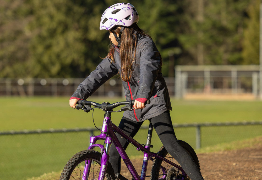 Kask rowerowy LAZER Nutz KinetiCore 2.0 Różowy dla Dzieci (rozmiar 50-56) park dziecko w kasku siedzi na rowerze patrzy w bok w tle trawnik drzewa bezpieczeństwo i komfort najmłodszych użytkowników chroni głowę podczas upadków i uderzeń lekki i wygodny w noszeniu regulowane paski siateczka przeciw owadom osłona skroni ochrona i komfort