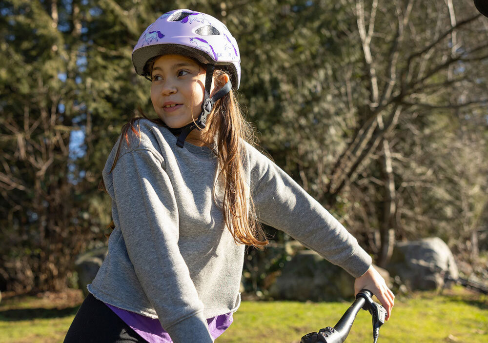 Kask rowerowy LAZER Nutz KinetiCore 2.0 Różowy dla Dzieci (rozmiar 50-56) park dziecko w kasku siedzi na rowerze patrzy w bok w tle trawnik drzewa klamra w kasku została sprytnie umieszczona na policzku eliminuje ryzyko bolesnego szczypania skóry podczas zapinania dzieci mogą łatwo i szybko dopasować kask komfort codziennego użytkowania pozytywne nastawienie do noszenia kasku wspiera bezpieczne poruszanie się