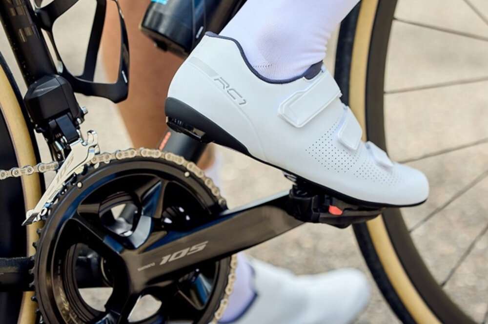 Buty rowerowe SHIMANO SH-RC102 Biały rozmiar 45 zbliżenie na korbę roweru osoba trzyma stopę opartą o pedał aktywność sport zdrowie