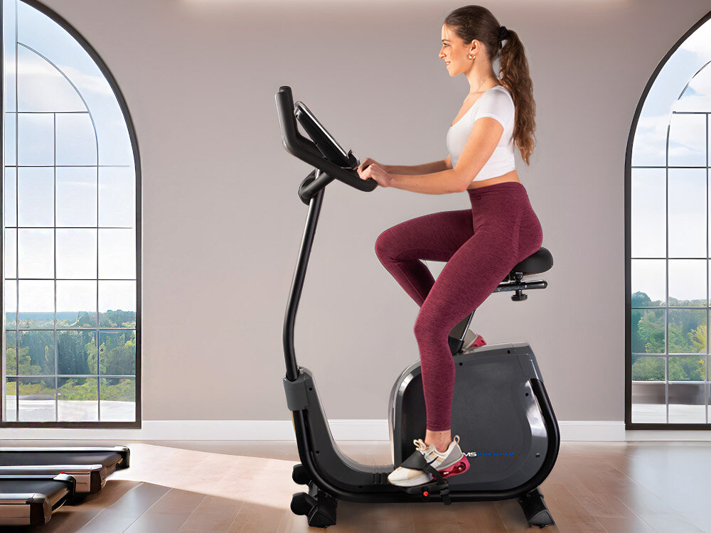 Rower elektromagnetyczny HMS PREMIUM M1815 kobieta trenuje na siłowni widok z boku inteligentny wyświetlacz LCD kontrolę kluczowych parametrów treningu ułatwia dopasowanie 19 programów 12 podstawowych 4 programy HRC programy celowe pomiar tętna odbywa się przez sensory na kierownicy uchwyt na tablet, kółka transportowe i niewielkie wymiary