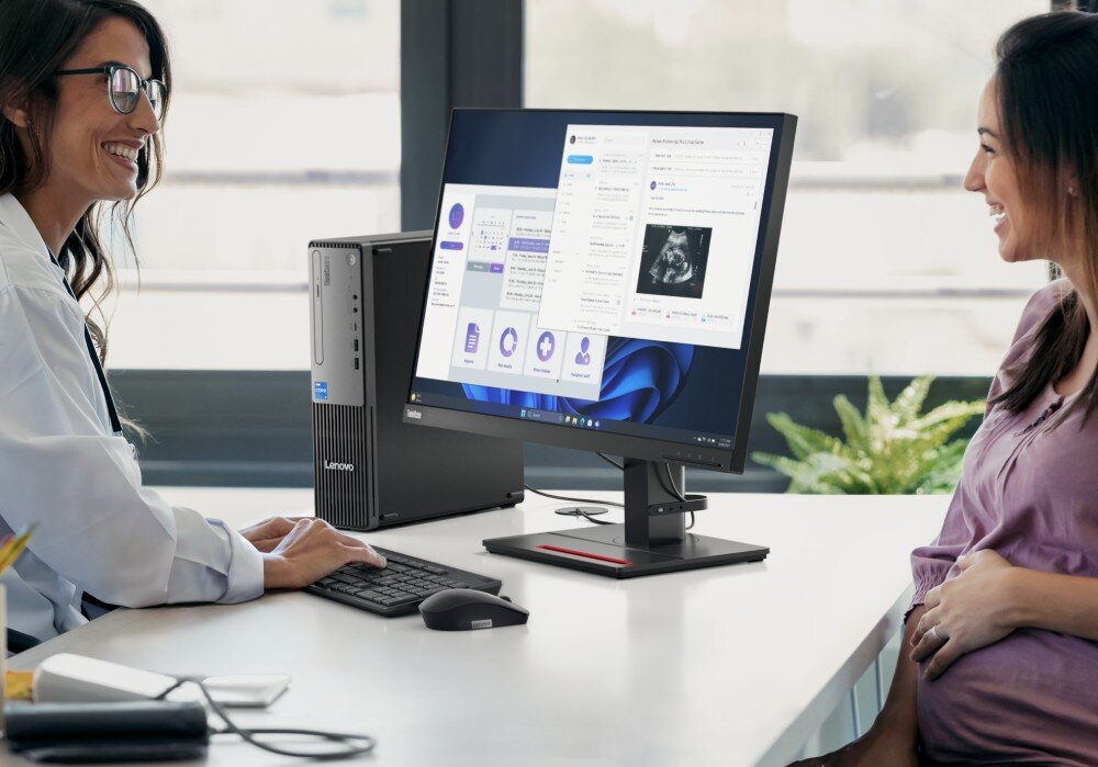Komputer LENOVO ThinkCentre Neo 50s Gen 5 Lekarz w białym fartuchu i okularach siedzi przy biurku i rozmawia z pacjentką, wskazując na monitor. Monitor spoczywa bezpośrednio na płasko położonej obudowie komputera, cechy, procesor, rdzenie, wątki