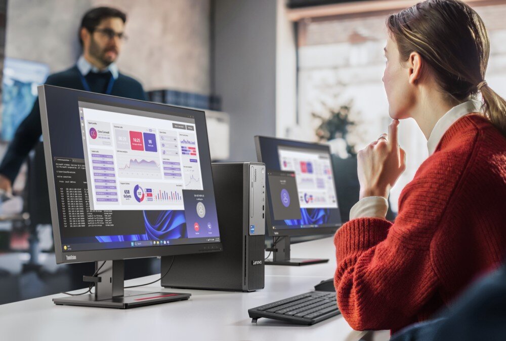 Na biurku znajdują się dwa monitory i komputer stacjonarny z widocznym logo Lenovo. Na ekranach wyświetlane są wykresy i dane, a w tle widać osobę stojącą.