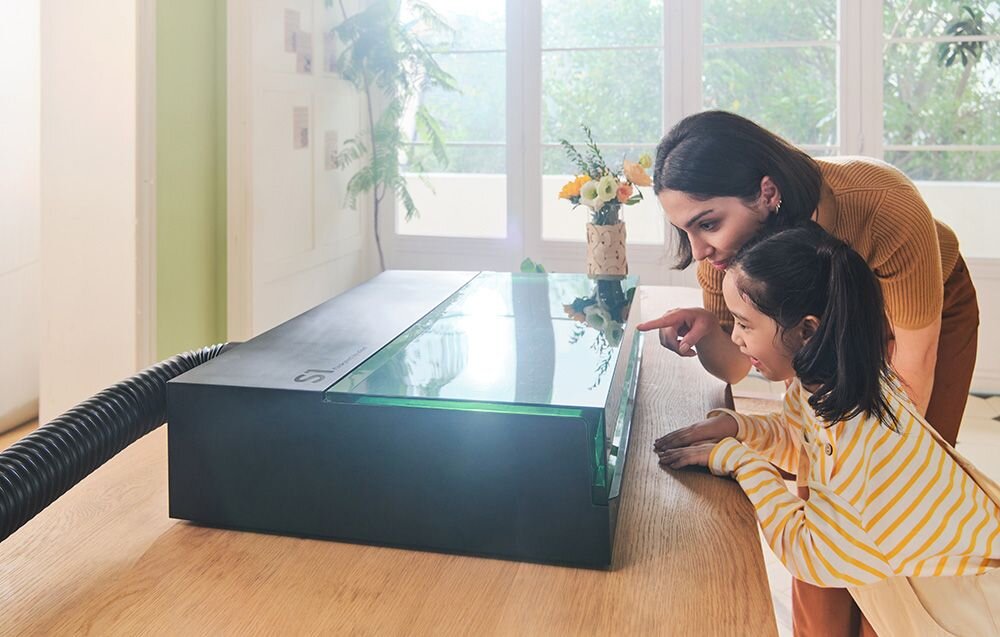 Grawerka laserowa XTOOL S1 40W (zestaw All-in-One) Kobieta i dziewczynka z ciekawością zaglądają przez szybę do wnętrza czarnego urządzenia na drewnianym stole.