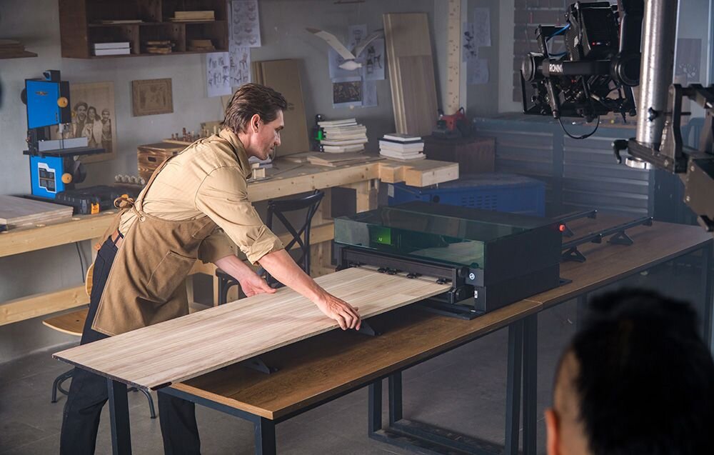 Grawerka laserowa XTOOL S1 40W (zestaw All-in-One) Mężczyzna w fartuchu wsuwa długą deskę do szerokiego urządzenia tnącego w warsztacie stolarskim cztery wymienne moduły przestrzeń robocza aplikacja xTool Creative Space