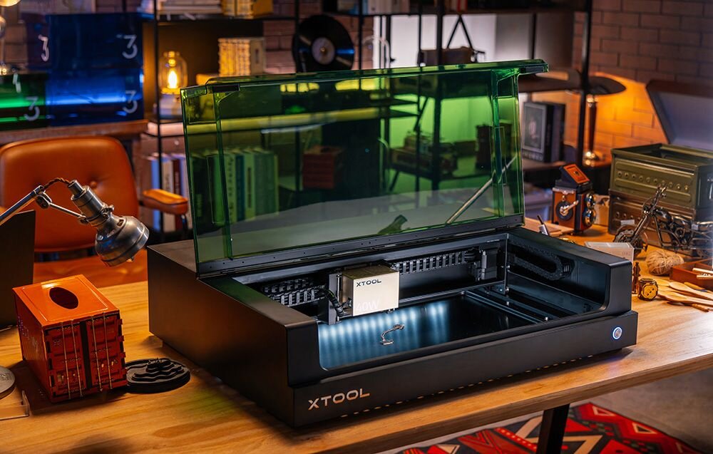 Grawerka laserowa XTOOL S1 40W (zestaw All-in-One) Otwarte czarne urządzenie z zieloną pokrywą stoi na biurku, ukazując ruchomą głowicę w środku oczyszczacz powietrza zdrowe środowisko trójwarstwowy filtr eliminacja szkodliwych cząsteczek