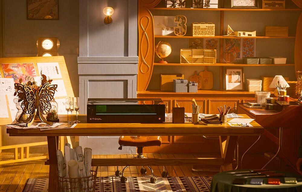 Grawerka laserowa XTOOL S1 40W (zestaw All-in-One) Czarne urządzenie stoi na środku dużego drewnianego stołu w jasnym, artystycznym pokoju z regałami regulacja przepływu powietrza stabilna energia ochrona soczewki poziom przepływu powietrza