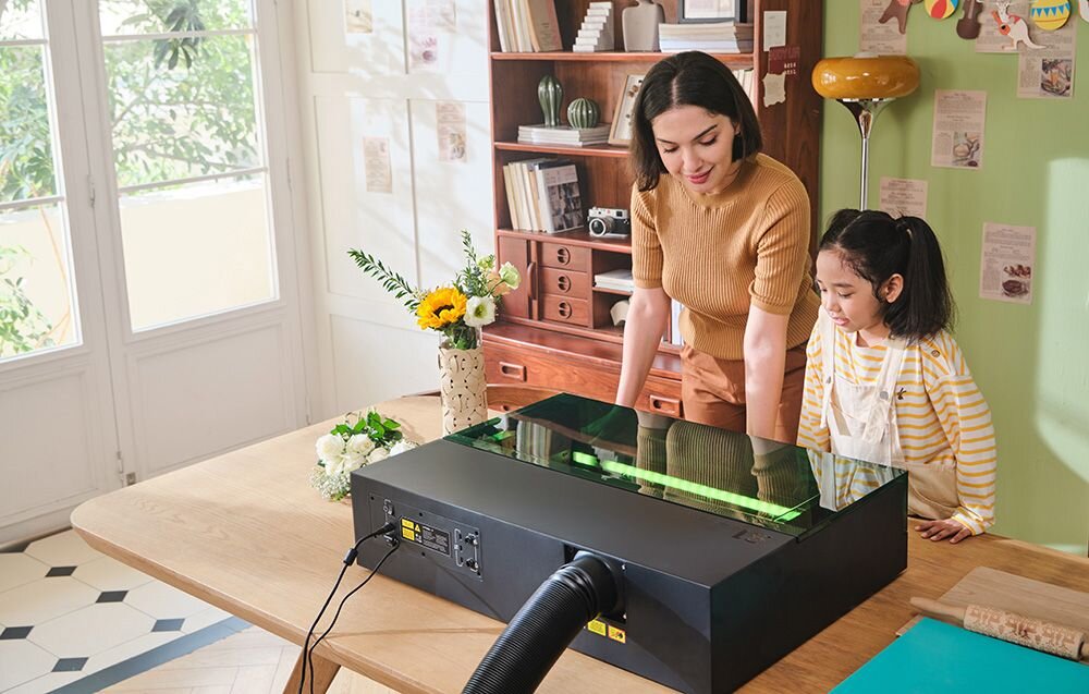 Grawerka laserowa XTOOL S1 40W (zestaw All-in-One) Kobieta i dziewczynka patrzą na pracujące urządzenie, z którego tyłu wychodzi gruba, czarna rura podstawa materiały o różnych grubościach regulacja płyty Riser Base