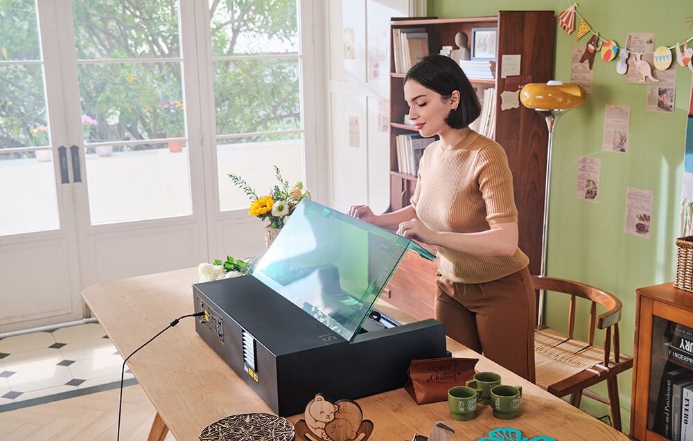 Grawerka laserowa XTOOL S1 40W (zestaw All-in-One) Uśmiechnięta kobieta unosi zieloną pokrywę urządzenia stojącego na stole obok kubków i dekoracji 4w1 możliwość regulacji w czterech różnych ustawieniach RA2 Pro