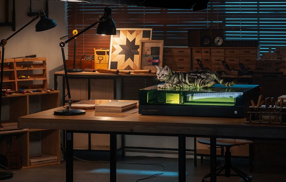 Grawerka laserowa XTOOL S1 40W (zestaw All-in-One) Kot siedzi na zamkniętej obudowie urządzenia w przytulnym, nastrojowo oświetlonym warsztacie system przeciwpożarowy ogranicza ryzyko wypadków