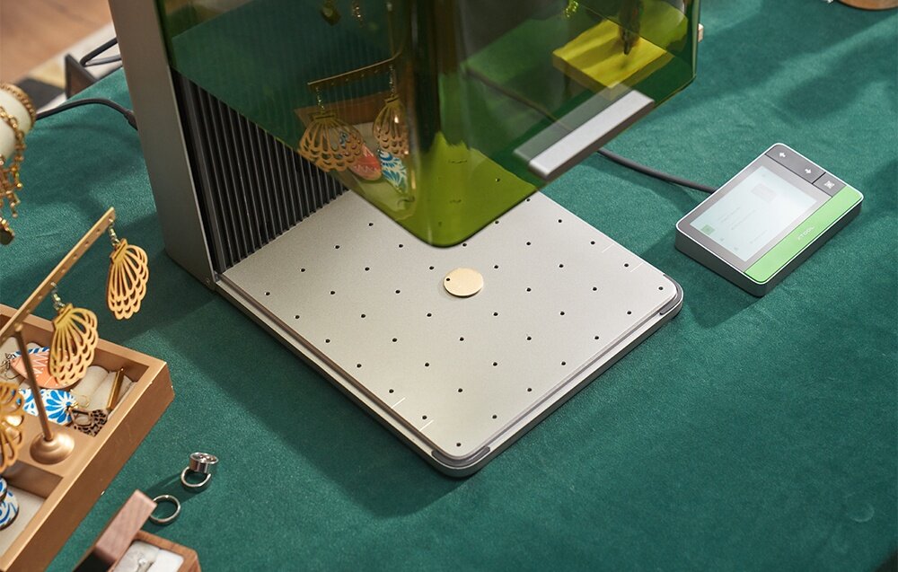 Na stole znajduje się grawerka laserowa z zieloną osłoną i metalową podstawą z otworami. Obok leży panel sterujący z wyświetlaczem, a w tle widoczne są ozdoby i akcesoria.