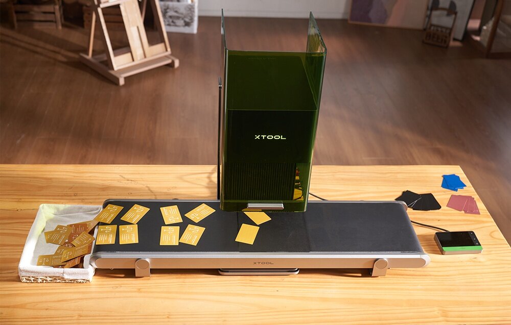Na drewnianym stole znajduje się grawerka laserowa z logo \'XTOOL\' oraz podajnik z kilkoma żółtymi kartami. Obok leży smartfon i kilka kolorowych próbek materiałów.
