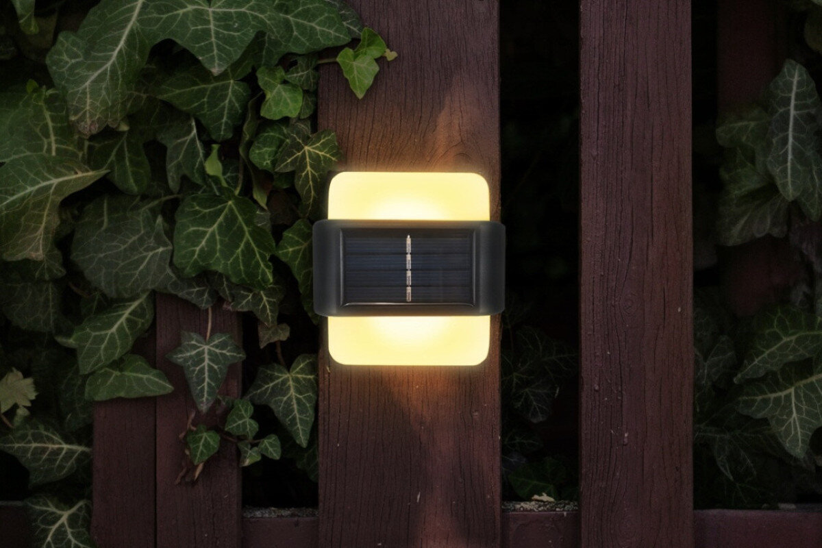 Kinkiet zewnętrzny solarny EXTRALINK Arras EX.24992 Czarny, ruch, czujnik, wejście, bezpiecznie, włączony kinkiet zamontowany do deski płotu, w tle rośliny