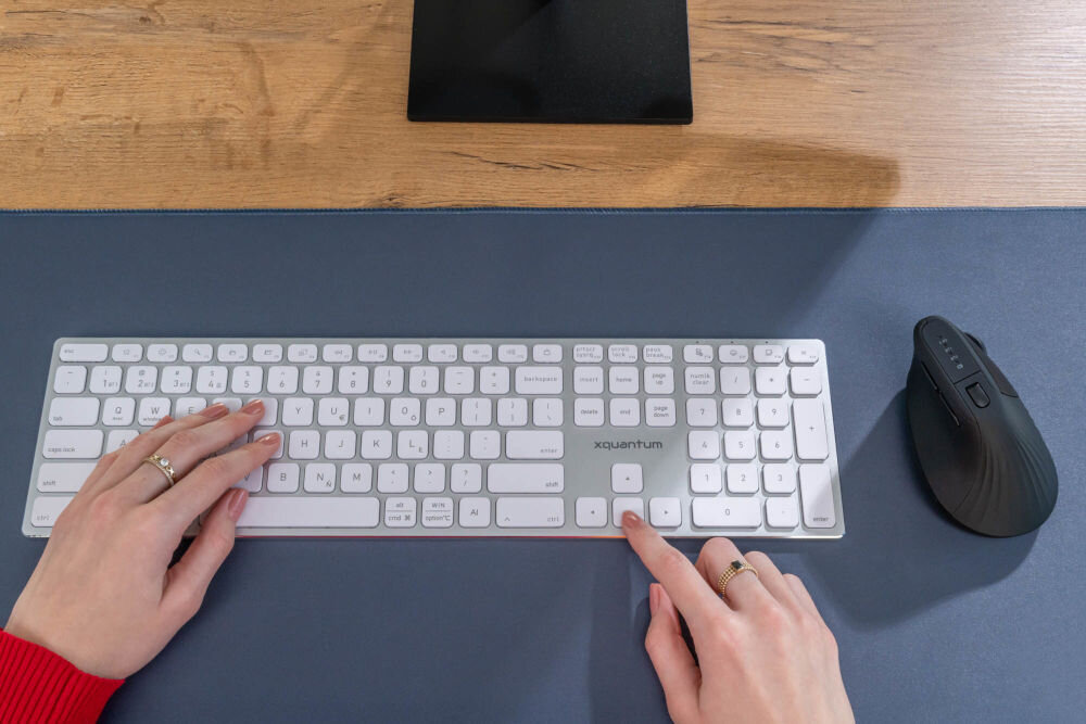 Mysz XQUANTUM WM700K Stanowisko pracy z białą klawiaturą XQUANTUM i czarną myszą ergonomiczną WM700K na niebieskim biurku.