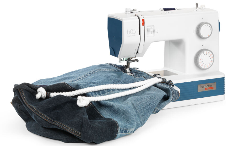 Maszyna do szycia BERNETTE 05 Academy Heavy Duty + Torba na maszynę maszyna stoi bokiem biało niebieska pokrętła suwaki szyty materiał jeansowy metalowa konstrukcja stabilność trwałość codzienne użytkowanie podczas intensywnej pracy 1100 wkłuć na minutę szybkie efektywne realizowanie projektów optymalne rezultaty w krótkim czasie