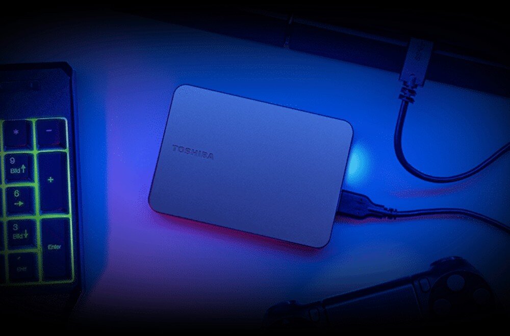 Dysk TOSHIBA Canvio Gaming Dysk zewnętrzny Toshiba na blacie, podłączony kablem USB C do góry, obok klawiatura numeryczna z podświetleniem i fragment kontrolera gier, opis