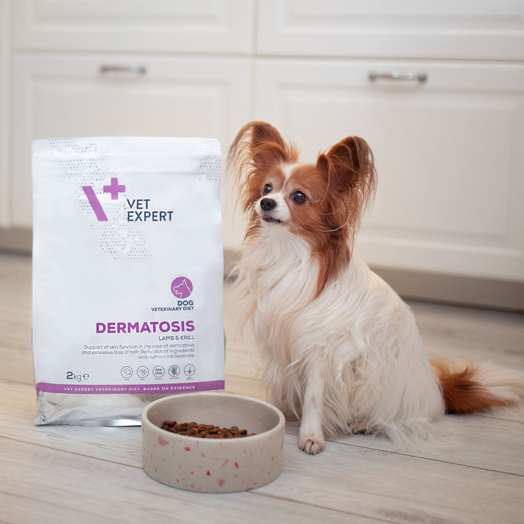 Na podłodze stoi worek z karmą \'Vet Expert Dermatosis Lamb & Krill\' o wadze 2 kg, obok którego znajduje się miska z karmą i pies. W tle widoczne są białe szafki kuchenne.