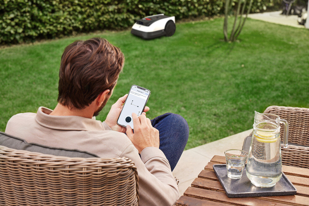 Robot koszący ECOVACS Goat O500 Panorama sterowanie Bluetooth-Wi-Fi osoba siedzi na krześle z telefonem w dłoni w tle robot na trawie aplikacja ECOVACS HOME Bluetooth Wi-Fi wygodne zarządzanie koszeniem z poziomu smartfona personalizowane plany koszenia dostosowywane harmonogramy pracy monitorowanie postępów w czasie rzeczywistym ręczne sterowanie robotem