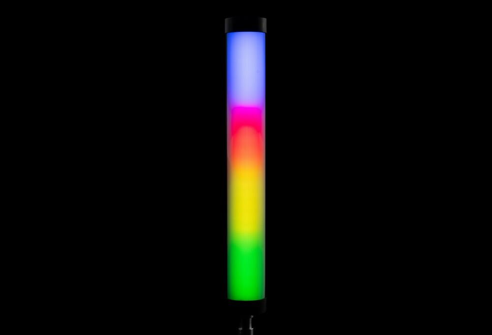 Lampa LED w kształcie tuby emituje wielokolorowe światło, widoczne są odcienie niebieskiego, różowego, czerwonego, żółtego i zielonego. Tło jest czarne, co podkreśla intensywność kolorów lampy.