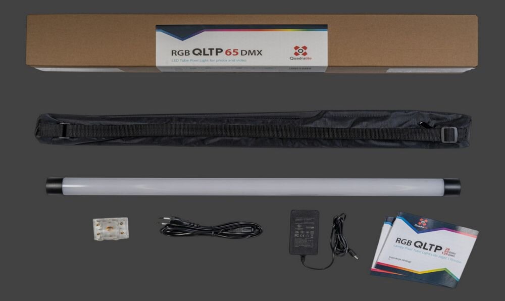 Na obrazie znajduje się zestaw z lampą LED w kształcie tuby, czarny pokrowiec, zasilacz, kabel oraz instrukcja. Na opakowaniu widoczny jest napis \'RGB QLTP 65 DMX\' oraz logo Quadralite.