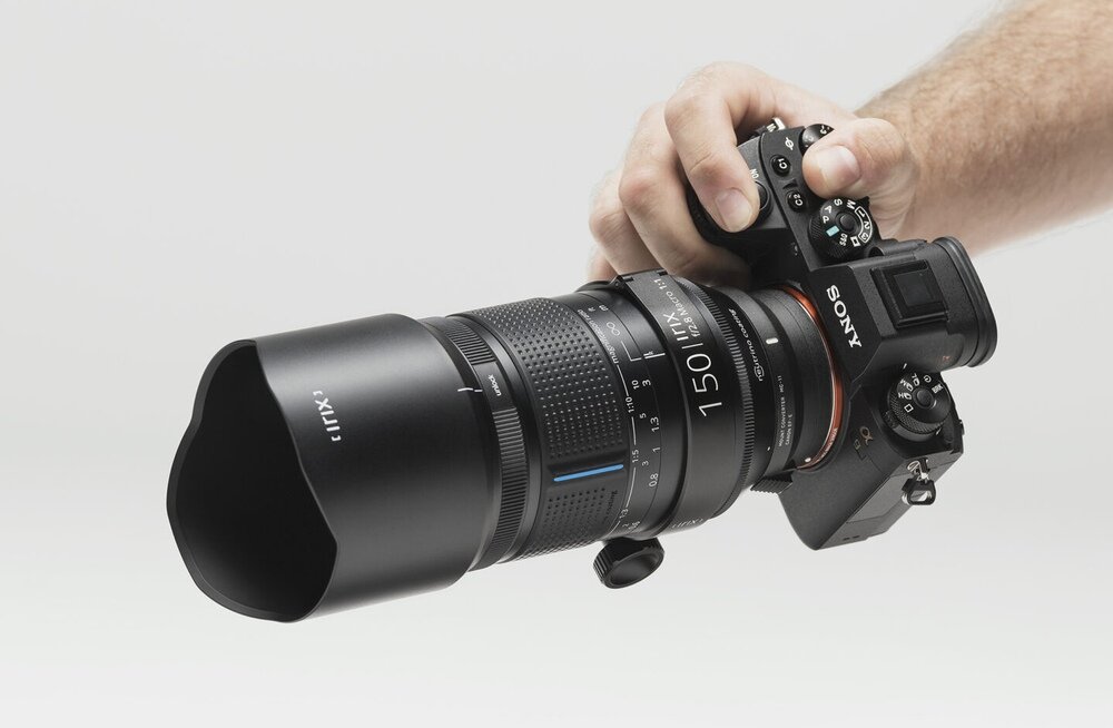Osoba trzyma aparat fotograficzny z zamontowanym obiektywem IRIX 150 mm f/2.8. Na obiektywie widoczne są napisy \'IRIX\' i \'150 mm f/2.8\'.