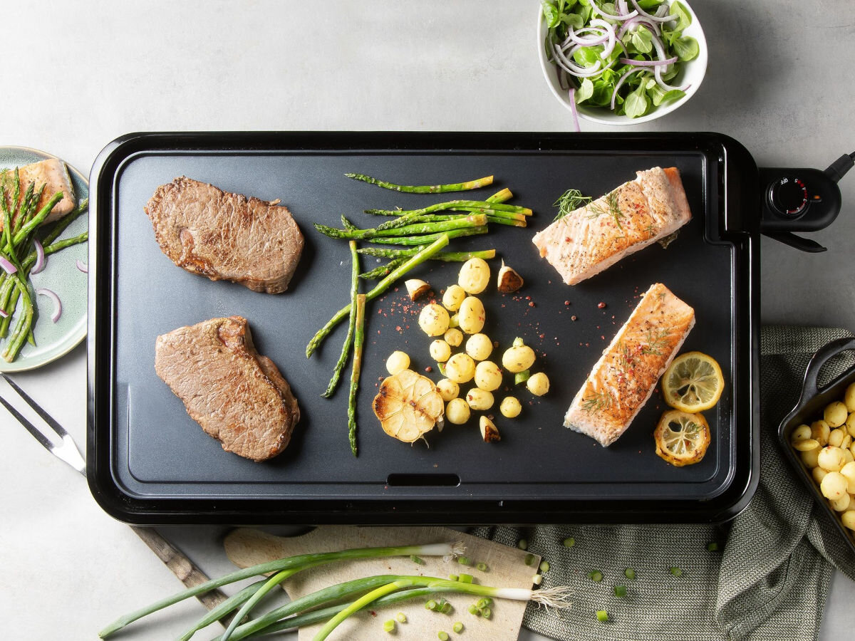 Grill DOMO Plancha XXL DO9263G, lekki grill, mobilność, łatwe przenoszenie, w domu i ogrodzie, Dwa steki, dwa filety łososia, zielone szparagi, ziemniaki i sałatka, grillowanie na płycie DOMO Plancha XXL, ujęcie lifestyle