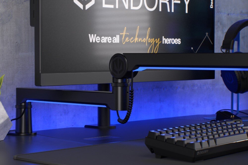 Ramię mikrofonowe ENDORFY Broadcast Low Profile RGB Boom Arm Ramię mikrofonowe ENDORFY Broadcast Low Profile Boom Arm Ramię zamocowane na biurku pod monitorem, obok klawiatury i słuchawek. Tło podświetlone niebieskim światłem, a na ekranie monitora widoczne logo producenta, przeznaczenie, zalety, cechy