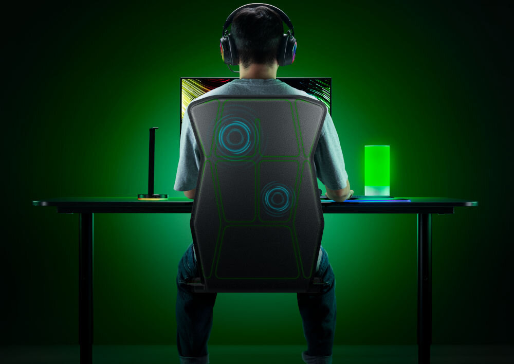 Mata haptyczna RAZER Freyja RC81-04340101-R3M1 prezentacja maty od tyłu i jej działania mężczyzna siedzący na niej przy komputerze w założonych słuchawkach na zielonym tle technologia haptyczna Razer Sensa HD dotykowe wrażenia intensywne eksplozje delikatne rytmy serca angażowanie realizm