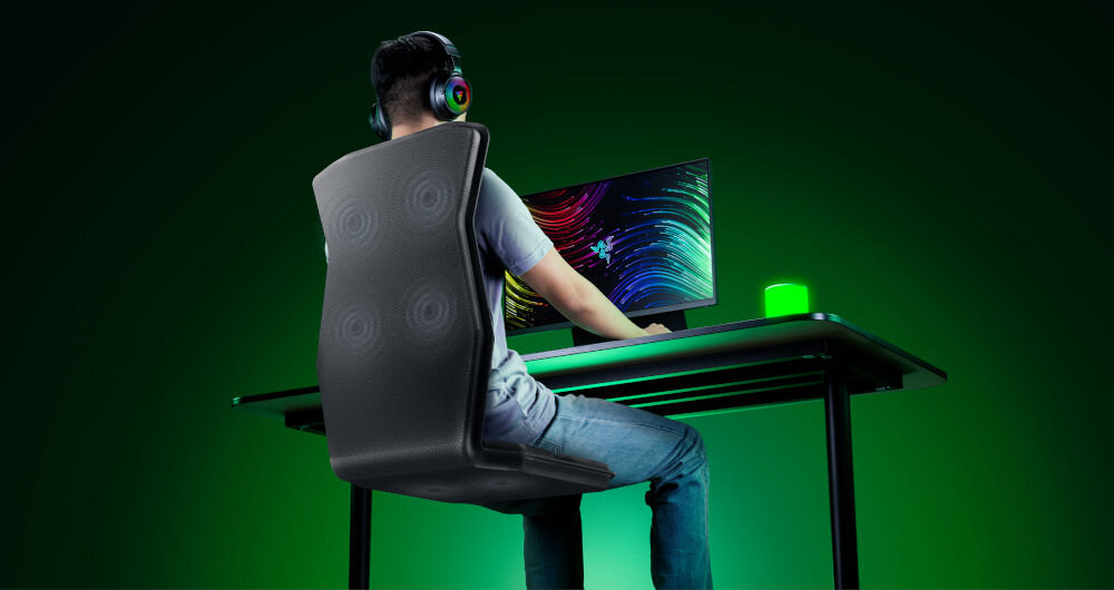 Mata haptyczna RAZER Freyja RC81-04340101-R3M1 prezentacja maty od tyłu i jej działania mężczyzna siedzący na niej przy komputerze w założonych słuchawkach na zielonym tle technologia haptyczna Razer Sensa HD wielowymiarowe wrażenia dotykowe odzwierciedlają intensywność kierunek czas trwania dźwięków realizm szczegółowość