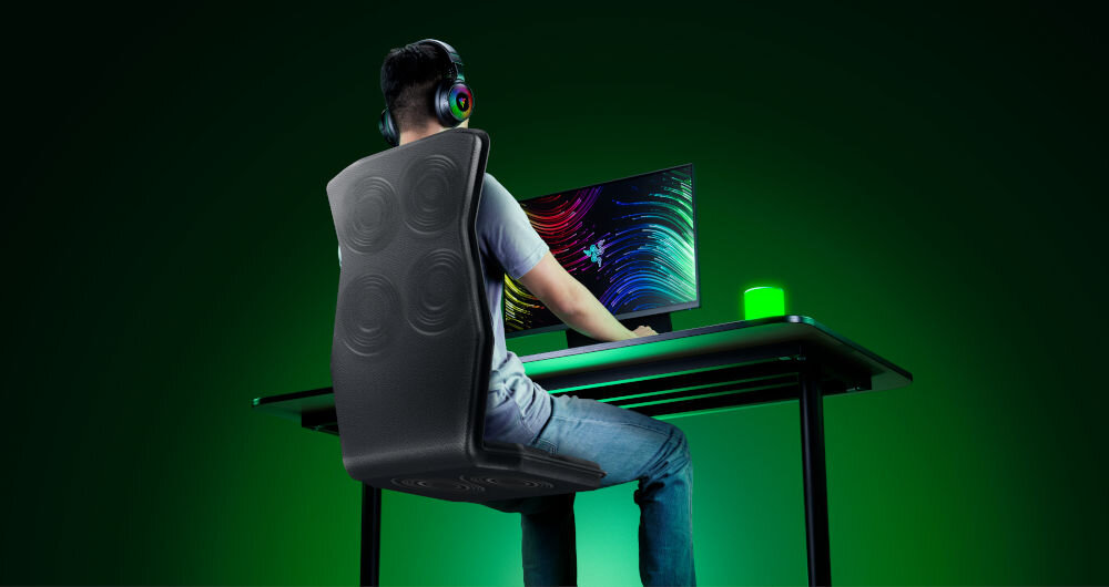 Mata haptyczna RAZER Freyja RC81-04340101-R3M1 prezentacja maty od tyłu pod kątem i jej działania mężczyzna siedzący na niej przy komputerze w założonych słuchawkach na zielonym tle sześć siłownikó haptycznych wrażenia dotykowe odczuwanie kierunku odległości i lokalizacji zdarzeń w czasie rzeczywistym immersja wyczuj dynamikę gry