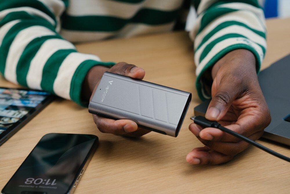 Dysk VERBATIM TurboMetal Mężczyzna trzymający urządzenie w jednej dłoni i w drugiej wtyczkę kabla, gotowy do podłączenia. Obok leżą smartfon i tablet, obudowa, materiał, chłodzenie, wentylator