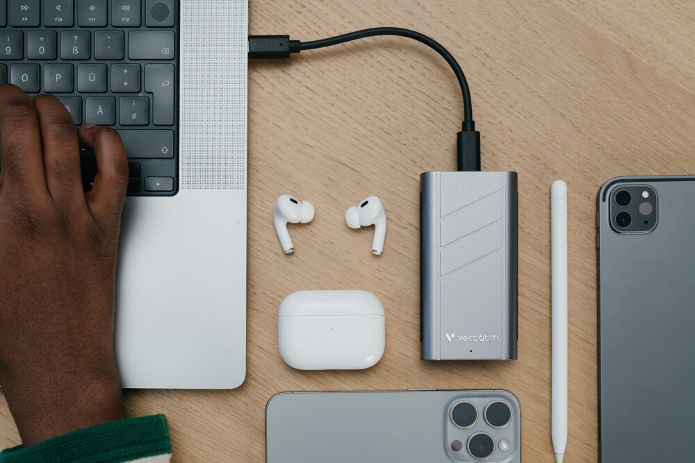 Dysk VERBATIM TurboMetal Widok z góry na urządzenie podłączone do laptopa. Wokół leżą słuchawki douszne, ich etui, rysik oraz dwa smartfony, oprogramowanie, kopie zapasowe
