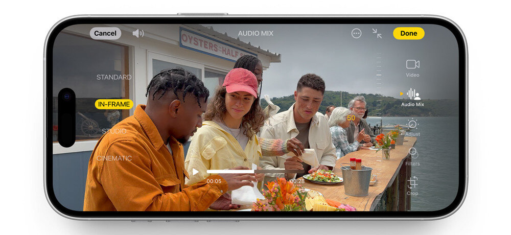 Na ekranie smartfona widać aplikację do edycji wideo z kilkoma osobami siedzącymi przy stole na zewnątrz. Widoczne są przyciski z tekstem: \'Cancel\', \'AUDIO MIX\', \'Done\', \'IN-FRAME\', \'STANDARD\', \'CINEMATIC\', \'Video\', \'Audio Mix\'.