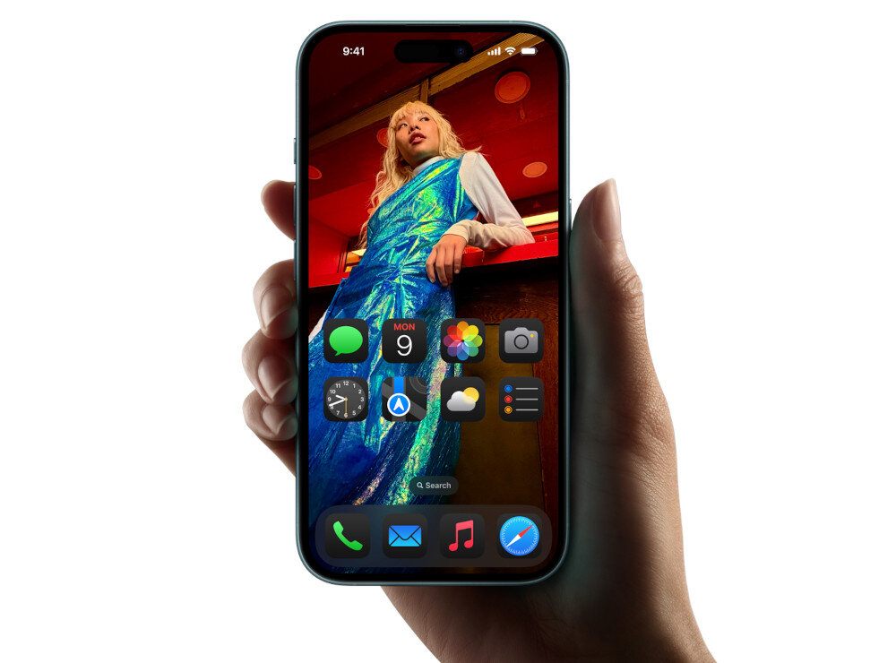 Osoba trzyma smartfon z wyświetlaczem pokazującym ekran główny z ikonami aplikacji. Na ekranie widoczna jest kolorowa fotografia osoby w niebieskiej sukience.