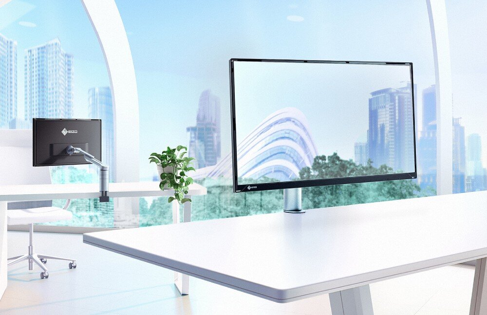 Monitor EIZO FlexScan FLT-BK Jasne, nowoczesne biuro z dużymi oknami. Na pierwszym planie monitor zamocowany na ramieniu do biurka. W tle drugi monitor i roślina doniczkowa na stoliku, opis