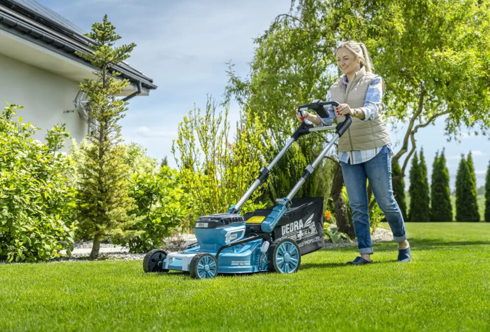 Kosiarka akumulatorowa DEDRA DED6998X z napędem ogród trawnik osoba kosi trawę w tle dom niebo drzewa kwiaty zadbany trawnik koszenie trawy mobilność zasilanie akumulatorowe