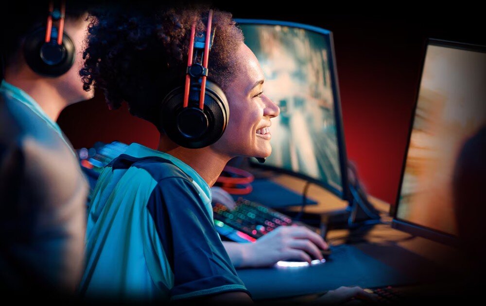 Karta graficzna SAPPHIRE Pure Radeon RX 9070 XT Gaming OC 16GB Kobieta w słuchawkach z mikrofonem uśmiechająca się podczas gry na komputerze, obok niej widać innego gracza i rozmyte monitory, technologie, funkcje