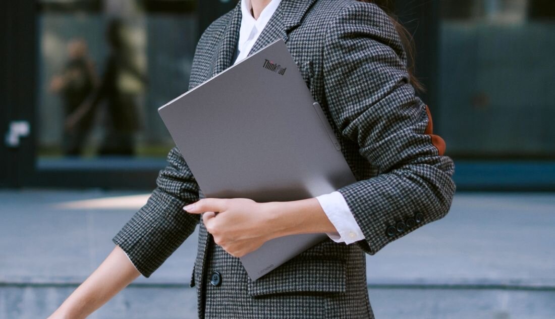 Osoba w garniturze niosąca szary laptop Lenovo ThinkPad w biznesowym otoczeniu.  